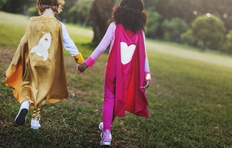 Two young children wearing superhero costumes with capes walking in the park and holding hands.